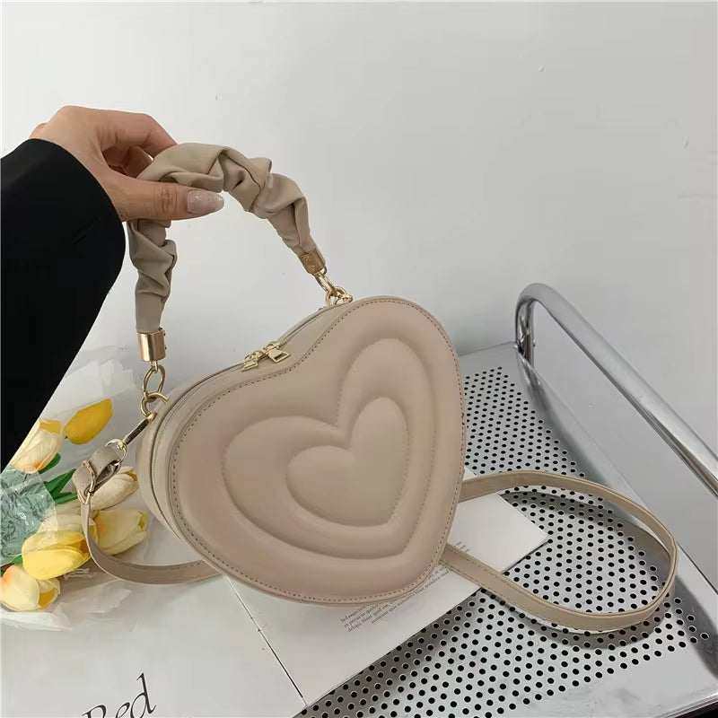 Heart-shaped shoulder bag in beige PU leather with top handle and crossbody strap.